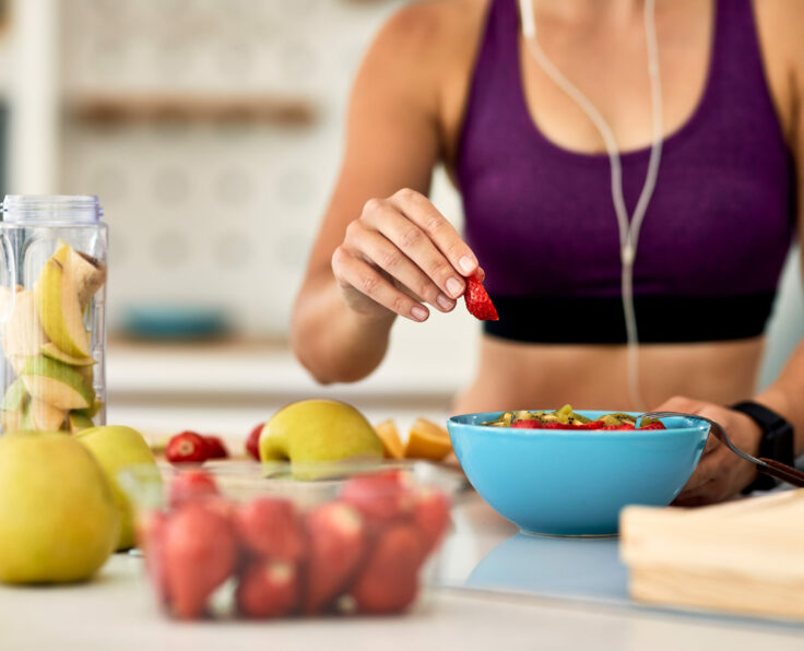 Close-up of athletic woman adding strawberries while making fruit salad in the kitchen.