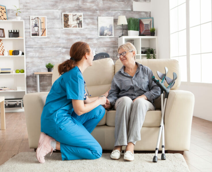 Happy old woman in a nursing home sitting on couch talking with her caretaker. Retired woman with crutches.